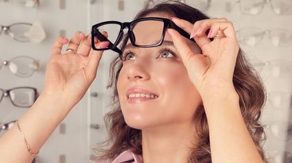 Lächelnde Person vor Wand mit Brillengestellen hebt Brille mit schwarzem Rahmen leicht vom Kopf ab und blickt hindurch