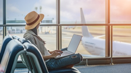 Person mit Brille und Hut sitzt mit einem Laptop im Wartebereich bereit zum Abflug