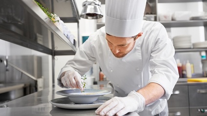 Person mit weißer Kochhaube und Arbeitskleidung, sowie Schutzhandschuhen bereitet eine Speise zu und steht dabei in einer Gastronomieküche