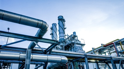 Im Vordergrund einer Industrieanlage verlaufen mehrere große metallene Rohrleitungen horizontal und diagonal durch das Bild. In der Bildmitte befinden sich weitere Rohrleitungen sowie Plattformen, Leitern und Geländer
