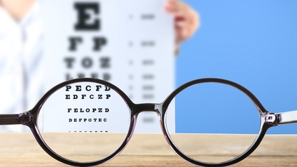 Brille mit schwarzem Rahmen im Fokus, auf Holzuntergrund liegend, durchscheinend aus blauem Hintergrund Sehtestplakat mit unterschiedlich groß verlaufenden Buchstaben