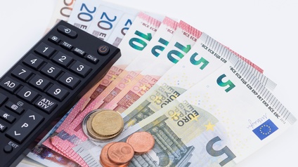 Eine Fernbedienung liegt auf nebeneinander aufgelegten Geldscheinen am Tisch, daneben sind noch ein paar Münzen gestapelt.