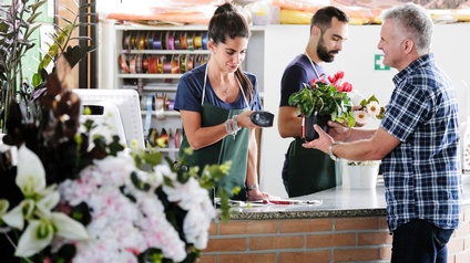 Person mit Blumentopf steht an Verkaufstheke eines Blumengeschäftes, zwei Personen mit Schürzen stehen dahinter, eine der Personen scannt Preisschild an Blumentopf, im Vordergrund verschwommen Blumen