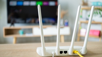 Auf einem Holztisch steht ein WLAN-Router, aus dem zwei Kabel kommen. Im Hintergrund ist ein großer Bildschirm