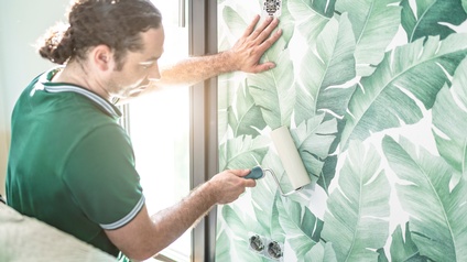 Person mit dunklen geschlossenen Haaren, Bart und grünem T-Shirt tapeziert eine Wand mit grünem Blättermuster