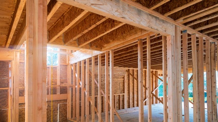 Rohbau eines Hauses, bestehend aus Holzstreben. Teilweise gibt es bereits Wände. Das Dach ist von innen verschlossen. In der linken Bildhälfte führt eine Treppe nach unten. Ein Stück darüber ist ein Fenster. Im Hintergrund ist eine grüne Landschaft