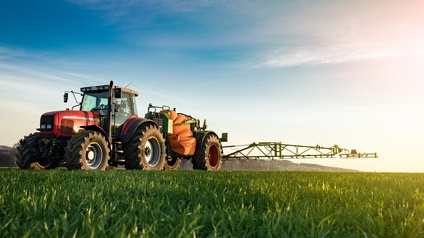 Roter Traktor mit Sprühvorrichtung fährt über grünes Feld unter blauem Himmel im Sonnenuntergang