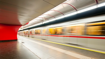 (KI-generierter Alttext) U-Bahn fährt mit Bewegungsunschärfe an einem leeren Bahnsteig in einem modernen Bahnhof mit roter Wand und Deckenbeleuchtung vorbei.