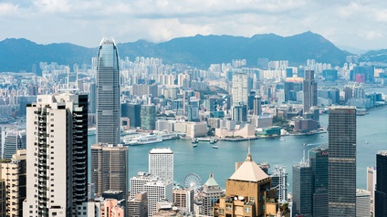 Stadtansicht von Hongkong: Blick von oben auf die am Wasser gelegene Stadt mit etlichen Wolkenkratzern unter bewölktem Himmel und Bergen im Hinterland. 