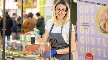 Porträt einer Person mit blonden schulterlangen Haaren, Brillen und Arbeitsschürze steht bei einem Stand und bereitet Speisen zu, während sie in die Kamera blickt