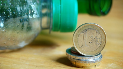 Ein Stapel Münzen mit einer aufgestellten 1 Euro-Münze vor einer Plastikflasche mit grünem Deckel und einer Grünglasflasche. 