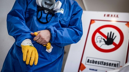 Mann säubert Gebäude von Asbest im blauen Schutzanzug mit gelben Handschuhen und einer Schutzmaske.  