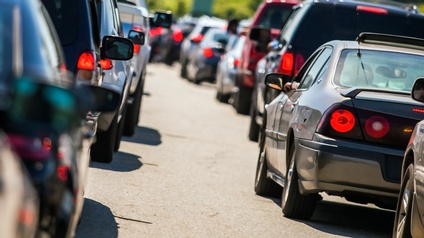Links und rechts stehen sehr viele Fahrzeuge sehr dicht hintereinander auf einer asphaltierten Straße. Die Lichter der Autos auf der Rückseite leuchten rot.