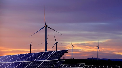 Im Vordergrund sind Solaranlagen auf einer Wiese. Dahinter sind Windräder im Licht der Dämmerung.
