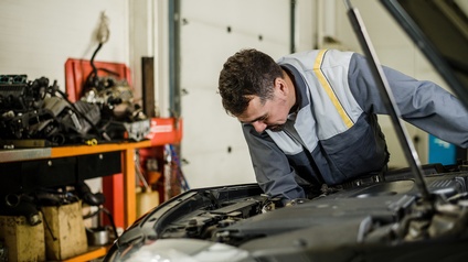 Person in Arbeitskluft in Werkstatt über Motor eines Autos gebeugt