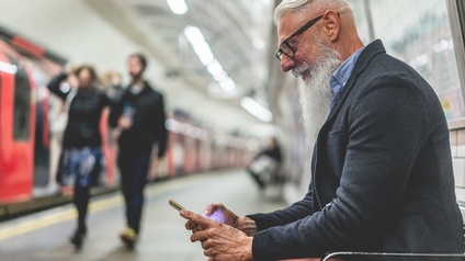 Lächelnde Person mit Bart und Brillen sitzt auf Sitzgelegenheit auf Bahnsteig und blickt auf Smartphone, im Hintergrund verschwommen weitere Personen und Zug