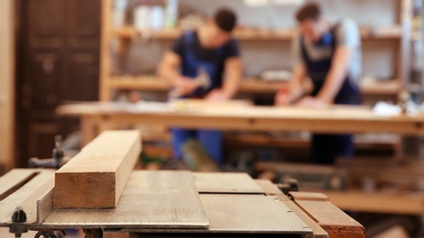 Im Vordergrund liegt ein heller, kleiner Holzbalken auf einer Holzplatte, die durch Schrauben an einer weiteren Platte montiert ist. Im Hintergrund verschwommen stehen zwei Personen an einem langen Holztisch. Die linke Person hält einen Hammer.