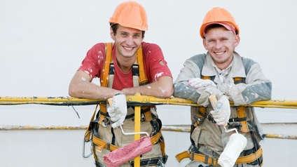 Zwei lächelnde Personen in Schutzbekleidungen lehnen an einem Gerüst und halten Malerrollen in der Hand