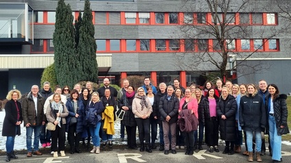 Eine größere Gruppe von Personen steht nebeneinander im Freien vor einem mehrstöckigen Gebäude. Die Personen tragen winterliche Kleidung und blicken in die Kamera. Einige halten Jacken oder Taschen in den Händen, alle stehen dicht beieinander.