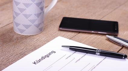 Nahaufnahme des oberen Teils eines Zettels, auf dem ganz oben Kündigung steht. Darunter sind schwarze Linien und stehen weitere Wörter. Auf und neben dem Blatt sind Stifte und liegt ein Smartphone.