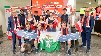 Gruppenbild von mehreren Personen, die teilweise Flaggen von Österreich sowie teilweise Schilder halten. Auf einem Schild steht Shanghai 8500 Kilometer. Auf einem anderen Schild steht Team Austria 2026