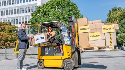Hella Riedl-Rabensteiner, Vorsitzende der Jungen Industrie Steiermark, sitzt auf einem Gabelstapler, Christian Wipfler, Landesvorsitzender der Jungen Wirtschaft, überreicht ihr ein Paket auf dem Netto geschrieben steht