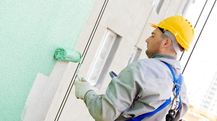 Person mit Schutzbekleidung und Sicherheitsgurt sowie Schutzhelm trägt mit Malerrolle grüne Farbe auf einer Hausfassade auf