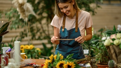 Lächelnde Person in Schürze blickt auf Sonnenblume vor ihr auf Tisch liegend und hält Strohfäden in Händen, ringsum verschwommen weitere Pflanzen
