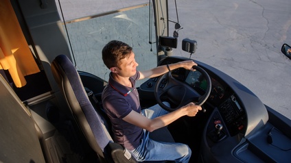 Eine Person mit Jeans und T-Shirt sitzt hinter einem Lenkrad eines Busses und hat beide Hände darauf