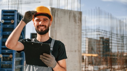 Porträt einer Person mit Helm, Handschuhen, Bart und Klemmbrett in der linken Hand. Hinter der Person sind mehrere Betonpflöcke und lange Metallstangen, die aus dem Boden ragen