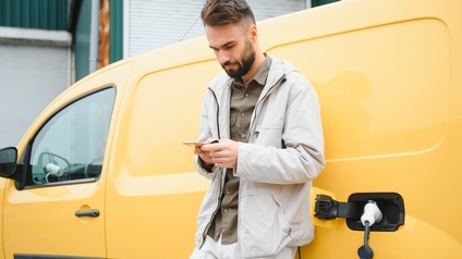 Ein gelbes Elektroauto wird mithilfe eines Ladekabels aufgeladen. Eine Person wartet währenddessen vor dem Auto. Sie ist an das Auto angelehnt und blickt auf ein Smartphone.