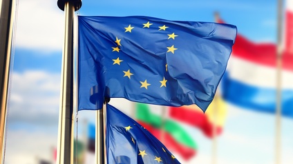 Im Vordergrund sind zwei Flaggen der europäischen Union auf Metallstangen, die im Wind wehen. Im Hintergrund sind weitere Fahnen auf Metallstangen, zum Beispiel die der Niederlande oder Spaniens