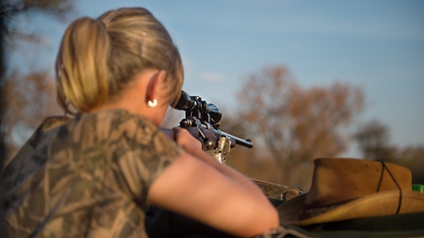 Eine Person ist aus der Rückenperspektive im Freien und hält ein Gewehr mit Zielfernrohr an der Schulter. Die Person trägt Kleidung mit Tarnmuster. Im Vordergrund befindet sich eine Auflagefläche, auf der das Gewehr abgestützt ist