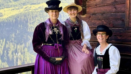 Drei Personen tragen traditionelle Trachten und stehen bzw. sitzen vor einer Holzhütte. Im Hintergrund sind Berge und Wald zu sehen. Alle drei lächeln in die Kamera, die Kleidung ist farblich aufeinander abgestimmt und detailreich verziert.