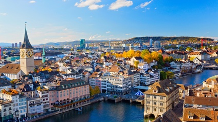 Panoramaaufnahme eines Flusses, an dessen Ufern links und rechts eine Stadt mit historischen Gebäuden ist. Im Hintergrund sind moderne Gebäude. Dahinter sind grüne Hügel