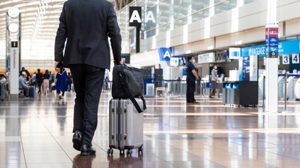 Rückenansicht einer Person in einem Anzug, die einen Rollkoffer mit einer Tasche darauf durch eine Halle eines Flughafens schiebt. Im Hintergrund sind verschiedene Schalter, Absperrungen und weitere Personen