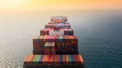 Frachtschiff auf offenem Gewässer fahrend in Vogelperspektive: Zahlreiche unterschiedliche färbige Container auf- und nebeneinandergestapelt, am Horizont verschwimmt Gewässer mit orangefarbenen Himmel