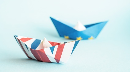 Zwei Papierschiffchen sind auf einem hellblauen Untergrund platziert, das vordere mit United Kingdom Flagge gefaltet und das hintere mit der Symbolik der Europäischen Union