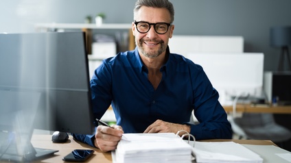 Porträt einer an einem Tisch sitzenden Person mit kurzen, grauen Haaren, Bart und Brille. Direkt vor der Person ist ein aufgeklappter mit einem Stapel Papier. Die Person hält einen Stift. Daneben ist ein Computer