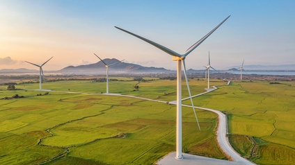 Landschaftsaufnahme einer grünen Landschaft, auf der versetzt Windräder stehen. Am Horizont ist eine Ortschaft vor einem Berg und neben einem Gewässer. Der Himmel ist blau und es herrscht Sonnenschein