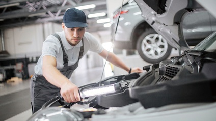 Person in Arbeitskleidung mit Kopfbedeckung lehnt sich über Motor eines Autos und blickt mit Leuchtstab hinein