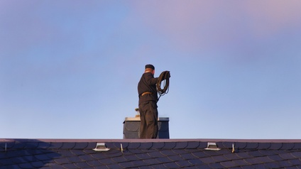 Person in Rückenansicht in schwarzer Arbeitskleidung mit schwarzer Kappe steht auf Dach vor Schornstein unter blauem Himmel und hält aufgewickeltes Drahtseil in Händen