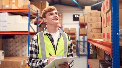 Lächelnde Person in gelber Schutzweste hält Stift und Clipboard in Händen und blickt vor Lagerregal stehend nach oben, im Hintergrund verschwommen weitere Regale gefüllt mit Kartonagen
