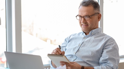 Person mittleren Alters mit schwarz-grauen kurzen Haaren, Brille und hellblauem Hemd sitzt freudig bei einem Schreibtisch, blickt auf einen Laptop und notiert sich etwas auf einem Notizblock