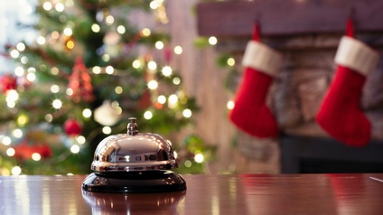 Eine Hotelklingel im Fokus, im Hintergrund verschwommen ein geschmückter Christbaum und Weihnachtsstrümpfe an einem Kamin hängend.