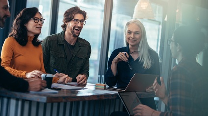 Personen stehen und sitzen freudig rund um einen Tisch bei einem Meeting mit Unterlagen und Laptops