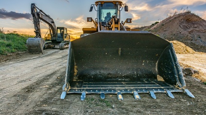 Ein gelbes Baufahrzeug mit einer großen Schaufel vorne steht auf einem erdigen Boden. Rechts davon ist ein Haufen Erde. Links davon steht ein Bagger. Neben dem Bagger ist grünes Gras. Im Hintergrund geht die Sonne am wolkenbedeckten Himmel unter