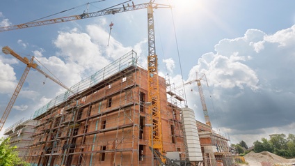 Frontalaufnahme eines Rohbaus eines Hauses mit Ziegelwänden. Um das Gebäude ist ein Gerüst aufgebaut. Daneben stehen mehrere Kräne