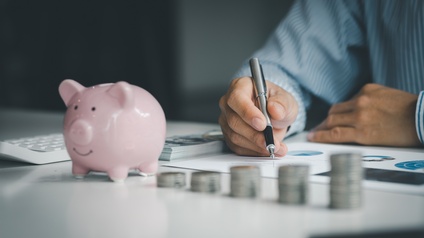 Auf einer Oberfläche sind fünf, unterschiedlich hohe Münzstapel. Links daneben steht eine kleine Figur eines Schweins. Links neben dem Schwein ist ein Taschenrechner. Dahinter sind die Arme einer Person. Unter der rechten Hand sind bedruckte Zettel