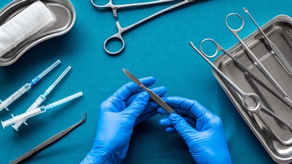 Auf einem türkischen Untergrund sind Werkzeuge für eine Operation platziert: ein Tuch, Spritzen, OP-Scheren, eine Pipette. Zwei Hände mit blauen Gummihandschuhen <br />
halten ein Skalpell. 